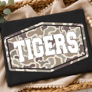 May include: A black t-shirt featuring a central hexagonal graphic with a camouflage pattern in shades of brown, tan, and grey. The word TIGERS is printed in large, bold, white block letters across the center of the camouflage hexagon design.