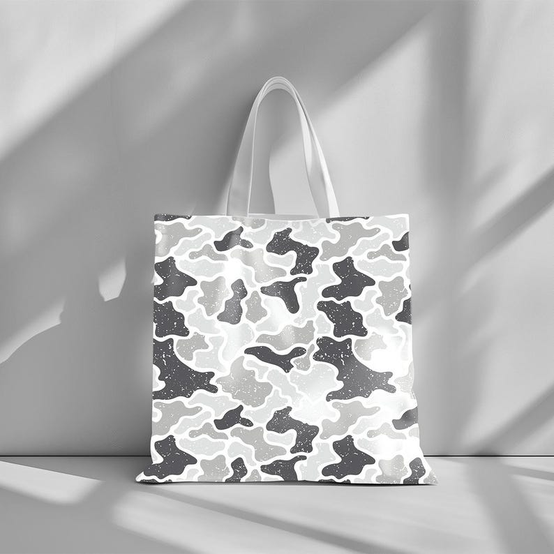 May include: A white tote bag with a repeating camouflage pattern in shades of gray. The bag has a simple design with a rectangular body and two white handles. The camouflage pattern has a distressed, textured look.