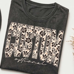 Op de afbeelding: Een antracietgrijs T-shirt met ronde hals en het woord CUSTOM in grote blokletters. De letters zijn gevuld met een beige, zwart en wit bloemen- en luipaardpatroon. Onder het woord staat het woord optional in witte schrijfletters.