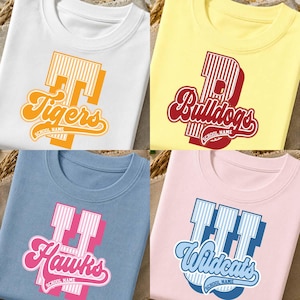 Pode incluir: Quatro camisetas de gola redonda nas cores branca, amarela, azul e rosa. Cada uma apresenta uma letra grande com listras verticais e uma sobreposição em fonte cursiva Tigers, Bulldogs, Hawks ou Wildcats, com SCHOOL NAME impresso abaixo.