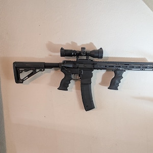 May include: Black rifle with a scope, magazine, and grip. The rifle is mounted on a white wall. The rifle has a black finish and a tactical design.