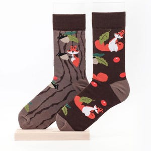 Könnte beinhalten: Ein Paar Socken mit Waldmotiv. Eine Socke zeigt ein braunes Baumstammdesign mit einem Fuchs, die andere einen dunkelbraunen Hintergrund mit Füchsen, roten Beeren und grünen Blättern. Beide Socken haben braune Bündchen.