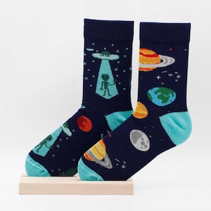 May include: A pair of blue socks with a space theme. The socks feature a green alien in a flying saucer, planets, and stars.