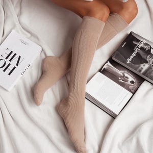 May include: A pair of beige knee-high socks with a cable knit pattern. The socks are worn by a person sitting on a white blanket.