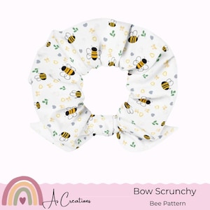 Puede incluir: Coletero blanco con un estampado de abejas. El coletero presenta un patrón repetido de abejas amarillas y negras, pequeñas hojas verdes y corazones grises. El coletero tiene forma de lazo y está diseñado para peinar el cabello.