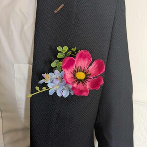 May include: A pink flower with a yellow center and blue flowers, with green leaves, pinned to a black suit jacket.