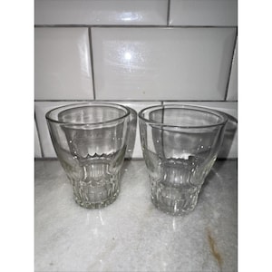 May include: Two clear, vintage-style drinking glasses. The glasses have a wide, rounded top and a detailed, textured base. The glasses are made of clear glass and are sitting on a marble-like surface.