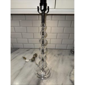 May include: A clear glass table lamp with a chrome stem and a series of clear glass spheres. The lamp has a round base and a black lamp holder. The lamp is plugged into an electrical outlet. The lamp is on a marble countertop.