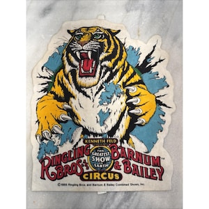 Peut inclure: Autocollant vintage du cirque Ringling Bros. et Barnum & Bailey représentant un tigre rugissant perçant un fond bleu. Le tigre est jaune et noir avec des yeux verts et une bouche rouge. Le texte "The Greatest Show on Earth" est inclus.
