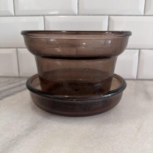 May include: Two brown glass bowls stacked on top of each other. The top bowl is slightly larger and has a flared rim. The bottom bowl is a shallow dish. The bowls are made of a transparent, dark brown glass.