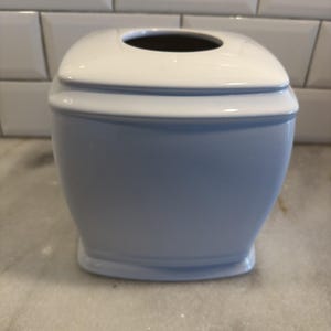May include: A square white tissue box cover with rounded corners and a smooth, glossy finish. The container features a slightly tapered body with a wide, centered circular opening at the top for dispensing tissues, showcasing a clean, solid design.