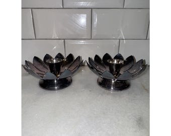 May include: Two silver lotus flower candle holders. Each holder has a central cup for a candle and petals that form the flower shape. The holders are on a white surface with a white tile background.