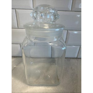 May include: A clear glass apothecary jar with a decorative lid. The jar is square-shaped with a rounded base and a clear glass lid with a knob. The jar is empty and sits on a light-colored surface.