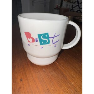 May include: White ceramic mug with a large handle and a stepped base. The mug features the words "BEST MOM" in colorful letters and a heart design. The mug is sitting on a wooden surface.
