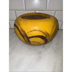 May include: A round, yellow ceramic planter with dark brown brushstroke designs. The planter has a wide opening and a textured surface. The interior is a darker brown color. The planter is sitting on a white and gray surface.