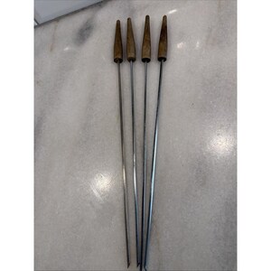 May include: Set of five metal skewers with wooden cone-shaped handles. The skewers are long and thin, with a silver finish. The wooden handles are a light brown color. These skewers are likely used for grilling or roasting.