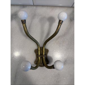 May include: A wall-mounted coat hook featuring a central brass-toned metal plate with an ornate embossed pattern. Four curved brass arms extend from the center, each ending in a smooth, round white ceramic knob for hanging garments or accessories.