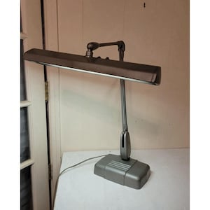 May include: A vintage gray desk lamp with a rectangular shade and a pivoting arm. The lamp has a rectangular base and a cord. The lamp is designed to provide focused light for reading or working.