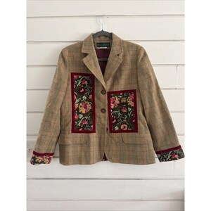 May include: A tan plaid blazer with floral embroidered patches and cuffs. The blazer has a dark red interior and trim around the floral patches. The blazer is hanging on a black hanger against a white background.