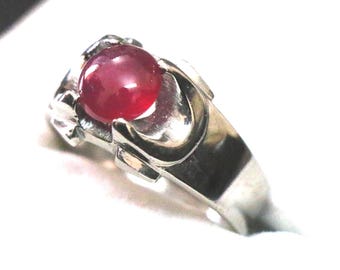 May include: A silver-toned metal ring featuring a smooth, round, deep red cabochon gemstone set within a pronged mounting. The band has a polished, reflective surface and a structural design that cradles the central stone securely in place.