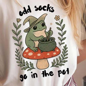 May include: White t-shirt featuring a cartoon frog wearing a witch hat, stirring a cauldron on a mushroom. The text "odd socks go in the pot" is printed above and below the frog illustration. The shirt has rolled-up sleeves.