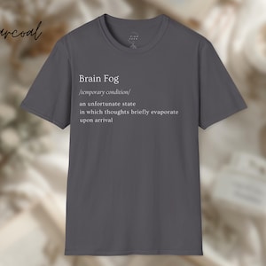 Puede incluir: Camiseta gris carb&oacute;n con la frase "Brain Fog" y una definici&oacute;n impresa en blanco. La definici&oacute;n describe una condici&oacute;n temporal en la que los pensamientos se evaporan brevemente. La camiseta es de cuello redondo.