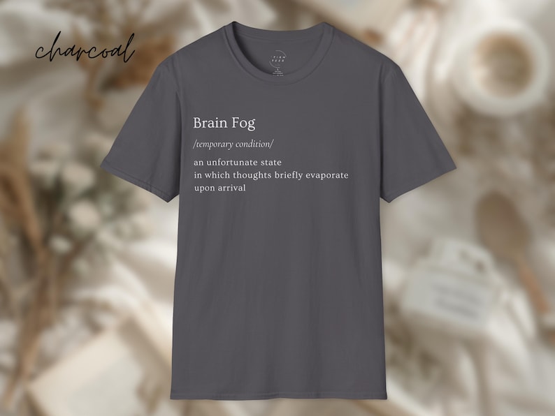 Puede incluir: Camiseta gris carb&oacute;n con la frase "Brain Fog" y una definici&oacute;n impresa en blanco. La definici&oacute;n describe una condici&oacute;n temporal en la que los pensamientos se evaporan brevemente. La camiseta es de cuello redondo.