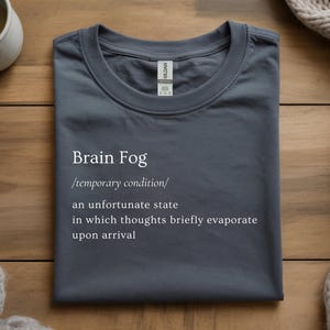 Puede incluir: Una camiseta gris oscuro con las palabras "Brain Fog" y una definici&oacute;n impresa en blanco. La definici&oacute;n describe una condici&oacute;n temporal donde los pensamientos se evaporan brevemente. La camiseta est&aacute; doblada sobre una superficie de madera.