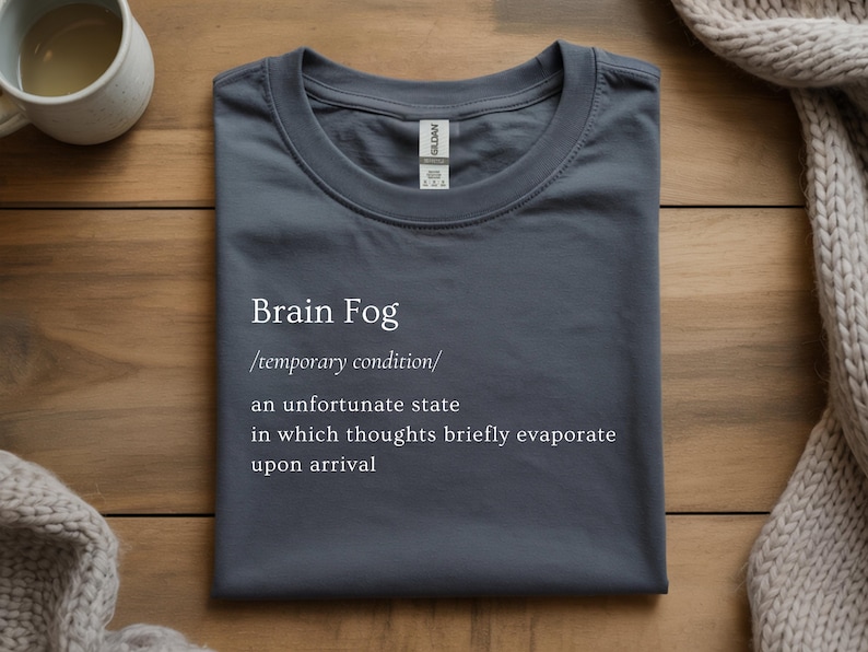 Puede incluir: Una camiseta gris oscuro con las palabras "Brain Fog" y una definici&oacute;n impresa en blanco. La definici&oacute;n describe una condici&oacute;n temporal donde los pensamientos se evaporan brevemente. La camiseta est&aacute; doblada sobre una superficie de madera.