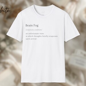 Puede incluir: Camiseta blanca con el texto "Brain Fog" y su definici&oacute;n. La definici&oacute;n describe un estado temporal en el que los pensamientos se evaporan brevemente. La palabra "white" est&aacute; escrita en cursiva en la esquina superior izquierda.