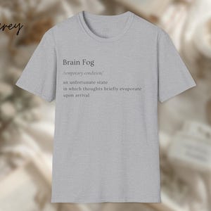 Puede incluir: Camiseta gris claro con la frase "Brain Fog" y su definici&oacute;n en texto negro. La definici&oacute;n describe una condici&oacute;n temporal donde los pensamientos se evaporan brevemente. La palabra "grey" est&aacute; escrita en cursiva en la esquina superior izquierda.