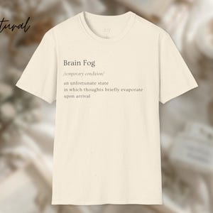 Puede incluir: Camiseta color crema con el texto "Brain Fog" y una definici&oacute;n. La definici&oacute;n describe una condici&oacute;n temporal donde los pensamientos se evaporan brevemente. La palabra "natural" est&aacute; escrita en cursiva.