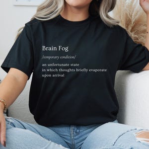 Puede incluir: Camiseta negra con la frase "Brain Fog" y su definici&oacute;n en blanco. La definici&oacute;n describe una condici&oacute;n temporal donde los pensamientos se evaporan brevemente. La camiseta se lleva con vaqueros azules.