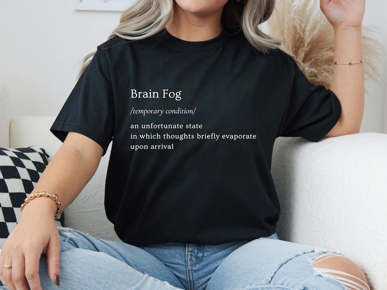 Puede incluir: Camiseta negra con la frase "Brain Fog" y su definici&oacute;n en blanco. La definici&oacute;n describe una condici&oacute;n temporal donde los pensamientos se evaporan brevemente. La camiseta se lleva con vaqueros azules.