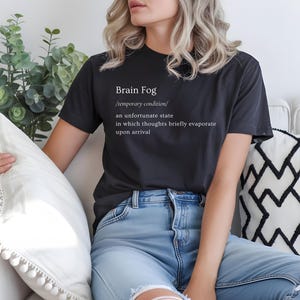 Puede incluir: Una camiseta gris oscuro con el texto "Brain Fog" y su definici&oacute;n en blanco. La persona lleva vaqueros azul claro. Est&aacute; sentada en un sof&aacute; blanco con cojines decorativos.