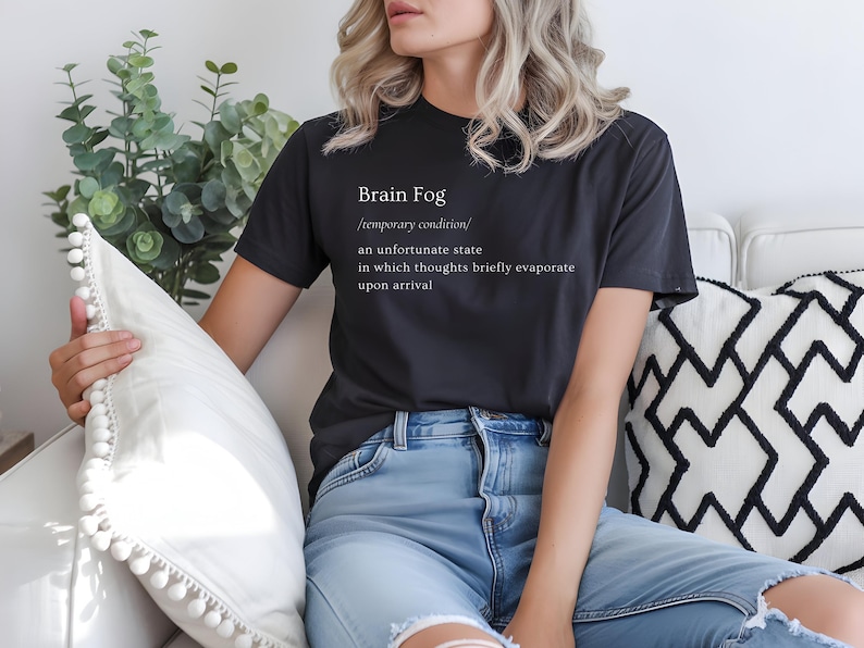 Puede incluir: Una camiseta gris oscuro con el texto "Brain Fog" y su definici&oacute;n en blanco. La persona lleva vaqueros azul claro. Est&aacute; sentada en un sof&aacute; blanco con cojines decorativos.
