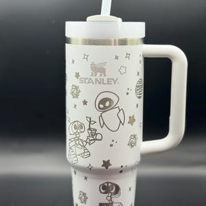 Peut inclure: Mug de voyage en acier inoxydable blanc avec une poignée noire et un couvercle blanc. Le mug a un fond blanc avec un motif noir et blanc des personnages du film Wall-E. Le mug porte également l'inscription "STANLEY".