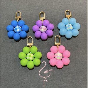 May include: Five flower-shaped keychains in various colors: blue, purple, light blue, green, and pink. Each keychain features a gold-colored clasp and a central bead with a plaid pattern. The flower petals are made of round, smooth beads.