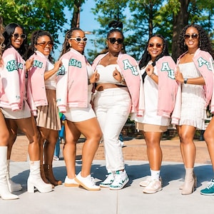 May include: A group of eight individuals wearing matching light pink and white varsity-style jackets featuring green and white embroidered patches that read AKA 1908. The jackets are worn over various white and beige tops, skirts, and pants.