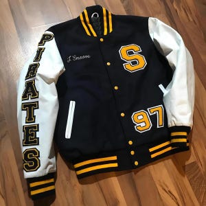 May include: Black and white varsity jacket with yellow accents. Features the word "PIRATES" on the left sleeve, a large yellow "S" and the number "97". The name "J. Smoove" is embroidered on the chest.