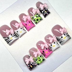 Pode incluir: Um conjunto de unhas postiças com vários designs. Algumas unhas apresentam um design de rosto de gato com um laço rosa, enquanto outras têm padrões rosa, verdes e pretos. Cada unha tem um laço rosa e uma ponta rosa claro.