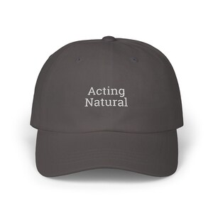 Peut inclure: Une casquette de baseball gris foncé avec un texte brodé blanc qui dit "Acting Natural".