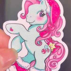 May include: A sticker featuring a cartoon pony with a light blue body, pink hair, and a Santa hat. The pony has pink cheeks, a heart on its side, and is wearing pink boots. The design includes candy and a star.
