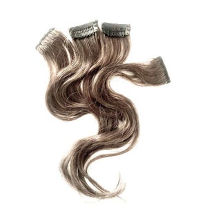 May include: A set of wavy hair extensions featuring a blend of dark brown and light ash blonde highlights. The hair strands are attached to three small, dark-colored metal snap clips at the top, allowing for secure attachment to natural hair.