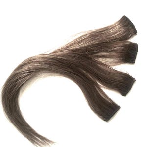 Puede incluir: Tres mechones de extensiones de cabello castaño con raíces oscuras y mechones castaños más claros. Las extensiones están dispuestas en forma curva sobre un fondo blanco. El cabello parece liso y lacio.