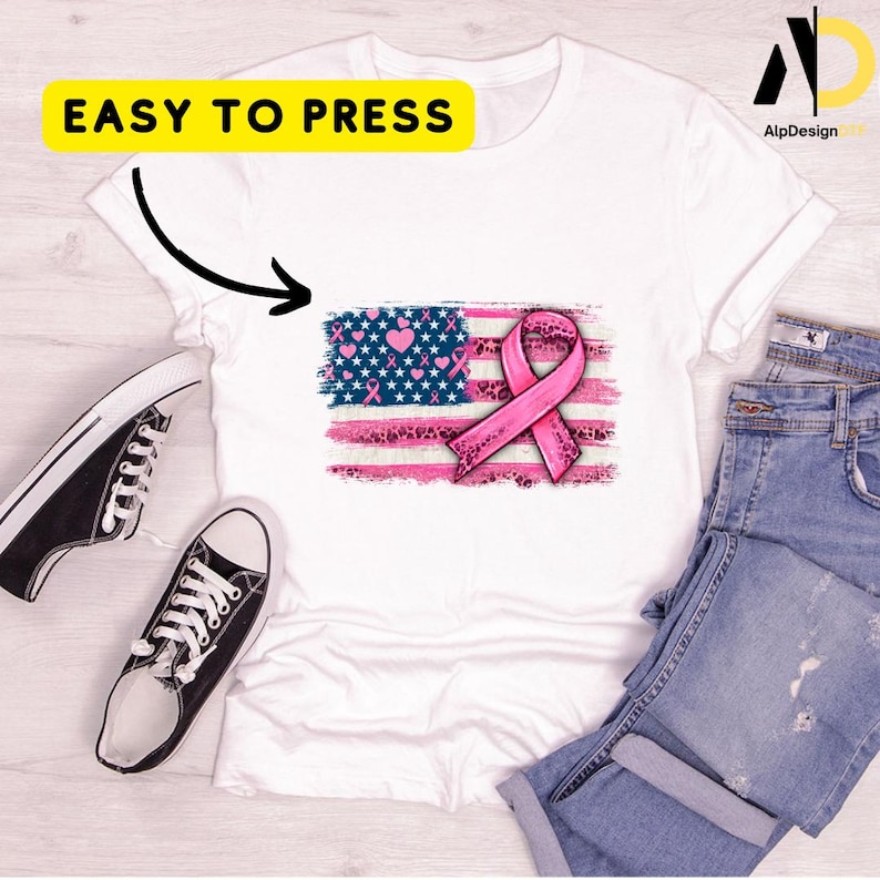 May include: White t-shirt featuring a pink ribbon and a distressed American flag design with hearts and stars. The text "EASY TO PRESS" is in a yellow rectangle. Black and white sneakers and blue jeans are also in the image.