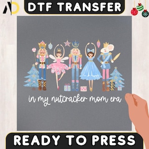 May include: A DTF transfer featuring a whimsical illustration of Nutcracker characters in shades of pink, blue, and gold, with Christmas trees and the text "in my nutcracker mom era". The design is ready to press.