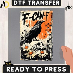 May include: A DTF transfer sheet featuring a graphic design with the text F-CAW-F. The illustration depicts a black crow perched on a human skull, surrounded by white daisies, small bats, and an orange circular background with a checkered pattern.