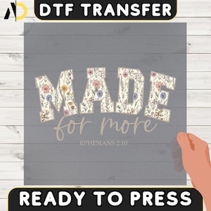 Peut inclure: Transfert DTF avec le texte "MADE for more" dans un motif floral, avec "Éphésiens 2:10" en dessous. Le transfert est prêt à presser, avec les mots "READY TO PRESS" en bas. Le fond est un grain de bois gris.