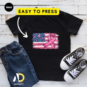 May include: Black t-shirt featuring a pink ribbon and an American flag design with hearts and stars. The words "EASY TO PRESS" are in a yellow rectangle above the design. A pair of black and white sneakers and blue jeans are next to the shirt.
